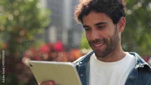 Portrait of handsome Hispanic man sends message on digital tablet in sunny city park. Young Latin American guy reads email as looks at mobile device in summer megapolis block