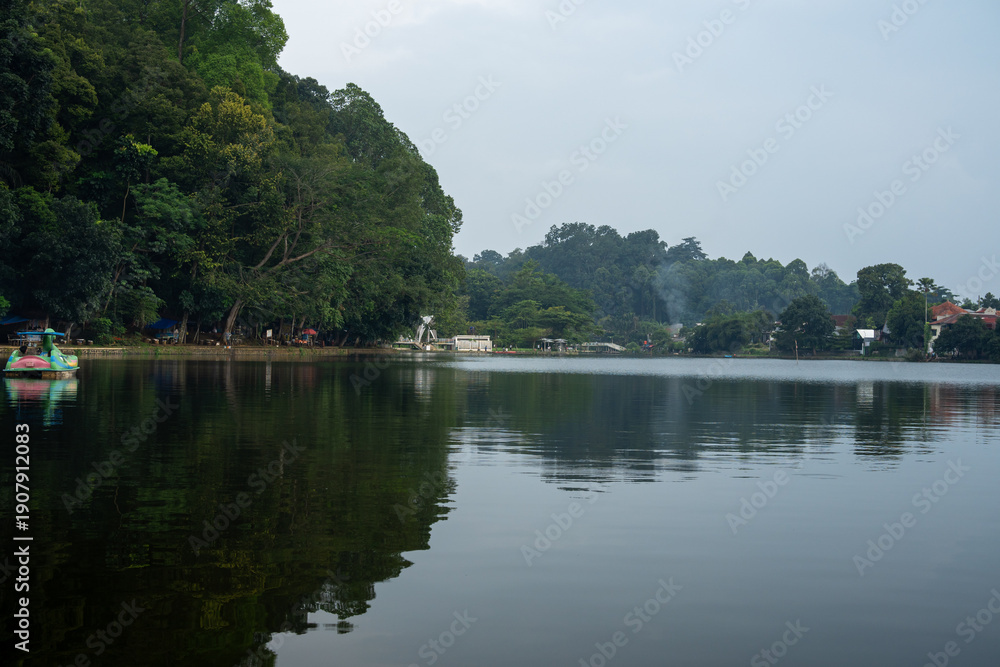 Fototapeta premium Calm lake surrounded by dense green forest and distant buildings
