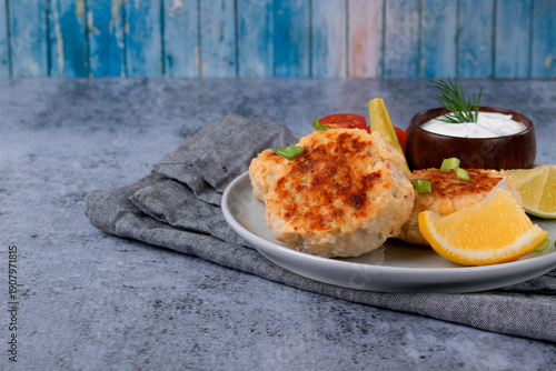 Fried fish cutlets made with bacon, cheese, and herbs are served with fresh vegetables, lemon wedges and sour cream sauce on a ceramic plate on a gray table