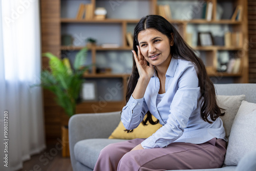 Konstfotografi Young indian woman sits on a sofa at home, pressing her ear with a pained expres