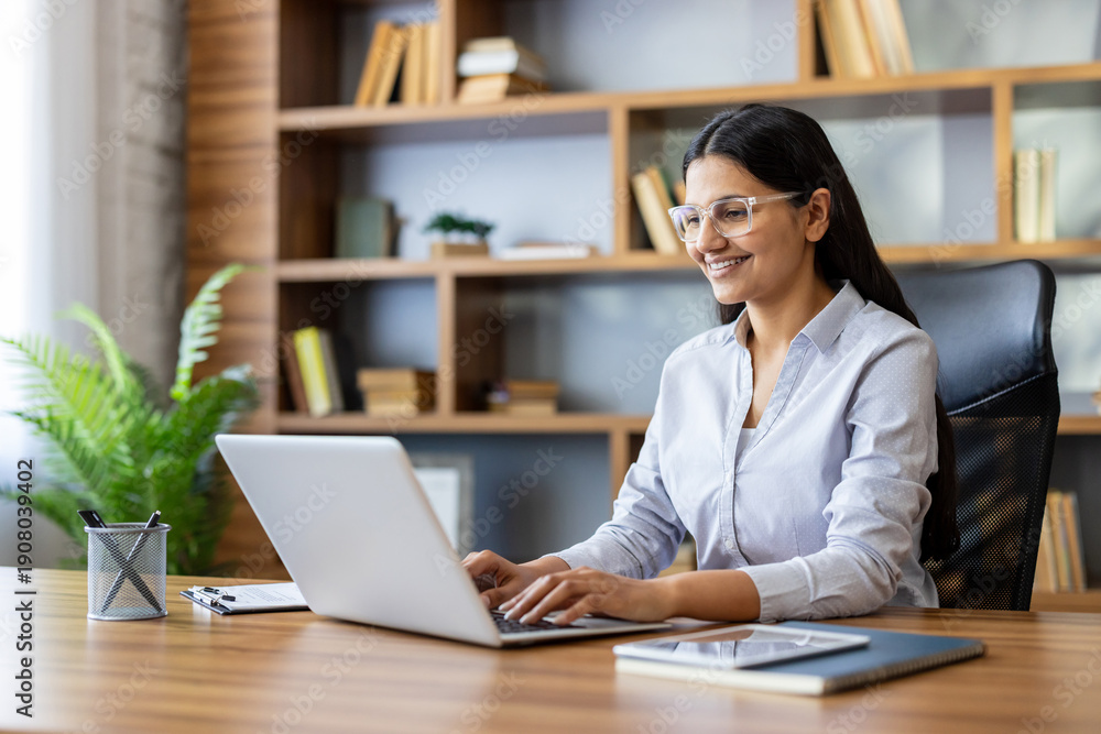 Fototapeta premium Young businesswoman with dark hair and glasses working on her laptop in a modern wooden office, smiling while typing, showing dedication and efficiency in her professional environment