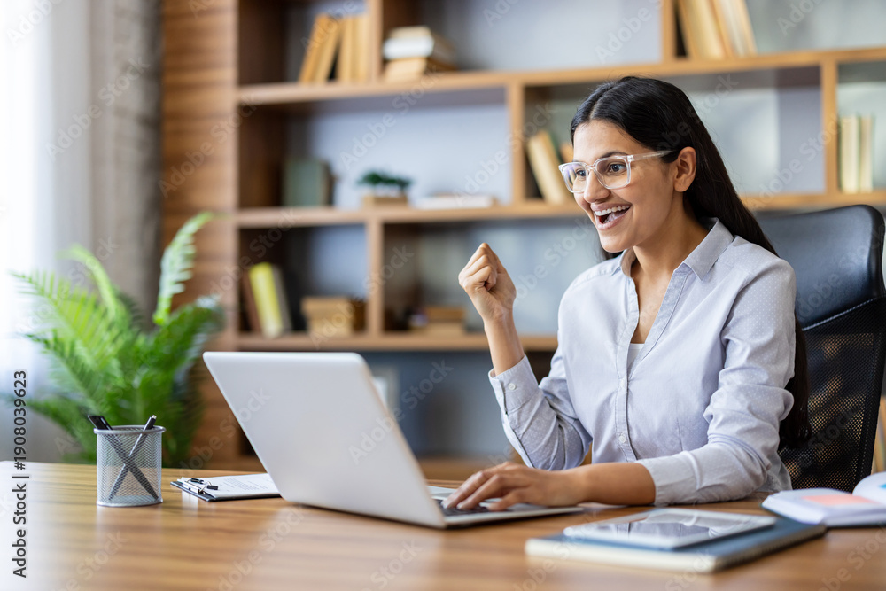 Fototapeta premium Indian woman at laptop pumps fist in celebration of career success and remote work achievement, smiling in a modern home office as a freelancer or entrepreneur completes a win