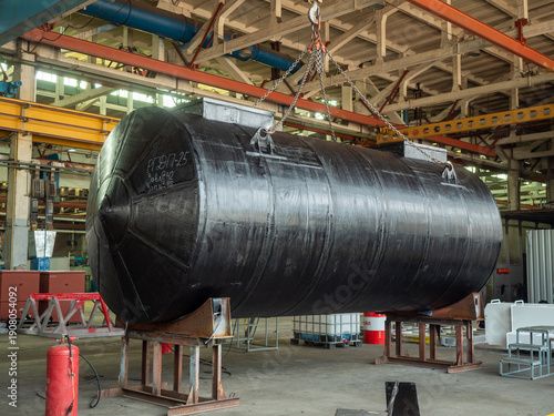 Foto Large horizontal black cylindrical metal storage tank suspended by heavy chains from overhead gantry crane inside industrial manufacturing facility workshop