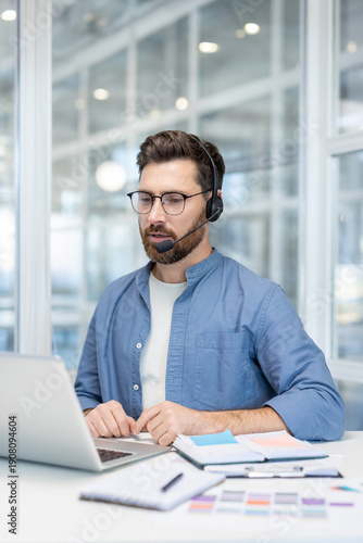 Male professional wearing a headset and spectacles, actively typing on a laptop while working on customer support, telemarketing, or online education in a modern office setup