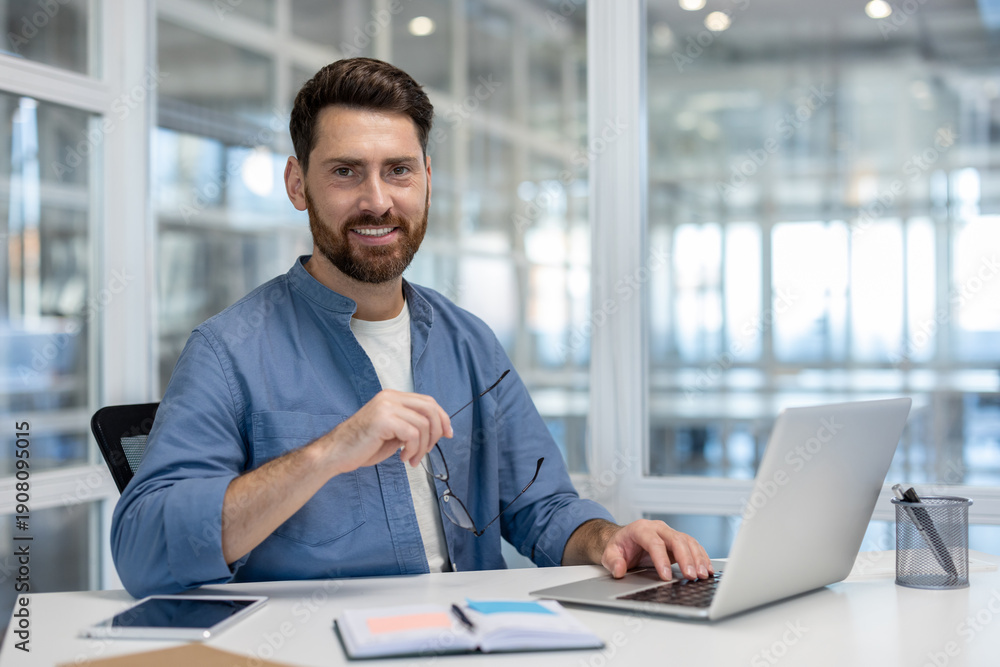 Fototapeta premium Confident bearded man smiling at the camera while working on a laptop at a bright desk, embodying professionalism, entrepreneurship, and productivity in a modern coworking space