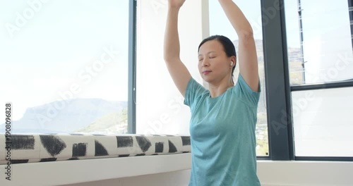 Asian woman sitting by windows on seat, earbuds guiding, joining palms into prayer, meditating