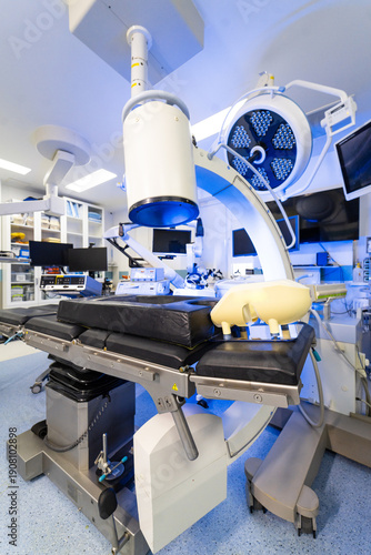 Modern hybrid operating room with robotic imaging technology. View of advanced medical equipment including C-arm and patient table in a sterile room.