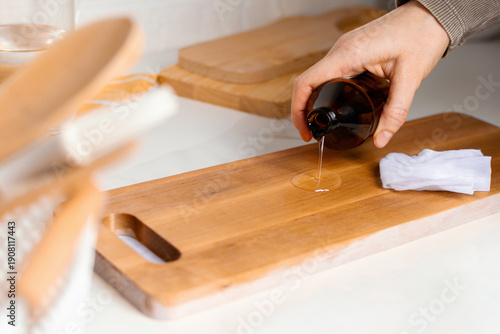 Hand pouring liquid oil from a dark glass bottle onto a wooden cutting board, with a cloth and kitchen utensils visible in the background, showcasing a clean and organized kitchen workspace.