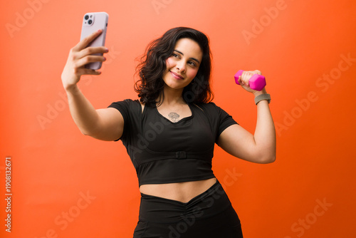 Fitness influencer woman taking selfie with smartphone and dumbbell, showing arm muscle in workout studio