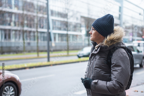 Close-up portrait of a man in a gray jacket with a black backpack waiting near a road with cars.