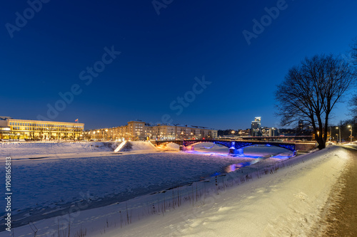 Night view of the frozen Neris River in Vilnius with Žvėrynas Bridge illuminated in red and blue lights, the Lithuanian Parliament in the background, and faint stars visible in the winter sky.