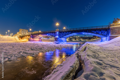 Night view of the frozen Neris River in Vilnius with Žvėrynas Bridge illuminated in red and blue lights, the Lithuanian Parliament in the background, and faint stars visible in the winter sky.