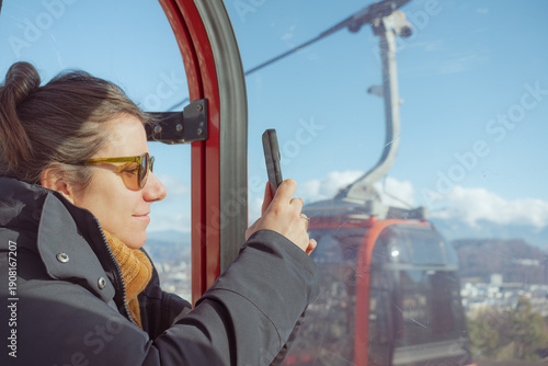 Bild auf Leinwand Young woman in a cable car gondola holding her smartphone to capture the panoram
