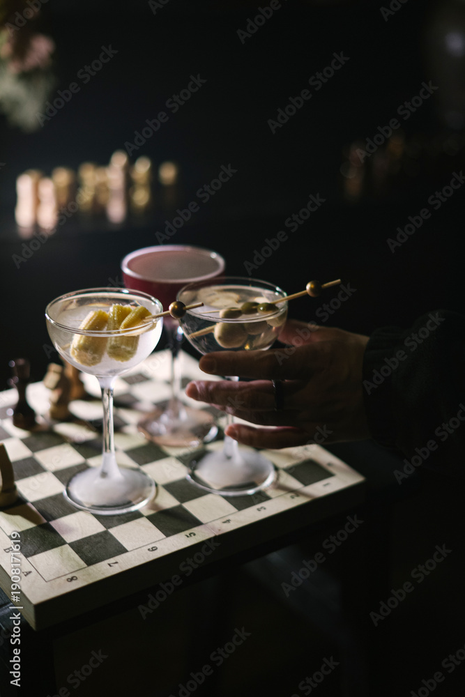 custom made wallpaper toronto digitalMan's hand holding a glass of dirty martini cocktail, with two more martinis standing on a wooden chess board in the dark room, close up view 