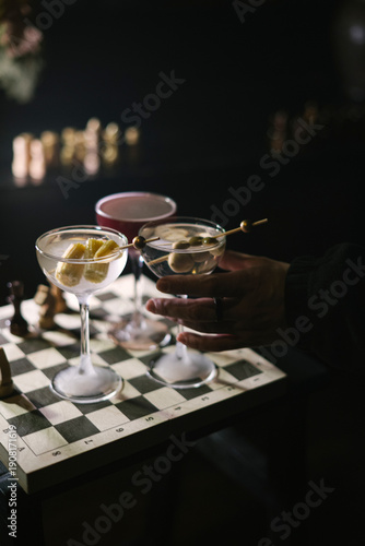 Wallpaper Mural Man's hand holding a glass of dirty martini cocktail, with two more martinis standing on a wooden chess board in the dark room, close up view  Torontodigital.ca