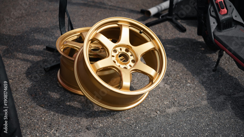 Pair of gold six-spoke alloy wheels on asphalt. Two aftermarket bronze forged rims resting on an asphalt surface, the primary wheel is angled toward the viewer, resting against a second identical one.