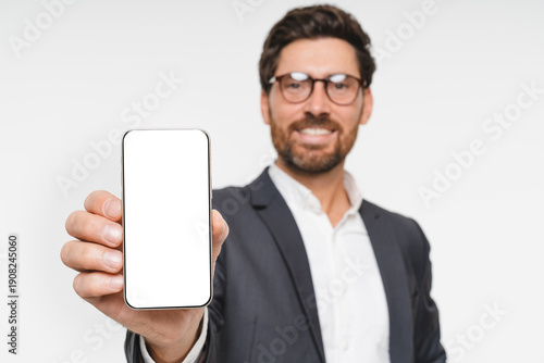 Smiling confident businessman showing mobile phone blank white screen mockup to camera standing isolated on background. Man in suit holding smartphone for finance business bank advertising app
