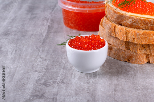 Red caviar in a small ceramic bowl on a gray table. Caviar sandwich in the background. Copy space