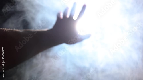 Close-up video with soft focus of a male hand reaching toward a powerful, voluminous projector light cutting through haze. Concept of striving for truth and enlightenment.
