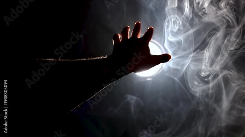 Video close-up with soft focus: a hand reaches into the frame toward a strong, volumetric light source shining through mist, representing a quest for enlightenment.