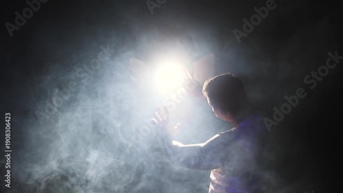 A symbolic close-up video. A man's hand stretches towards a luminous, powerful light cutting through smoky haze, evoking themes of faith, epiphany, and higher truth.