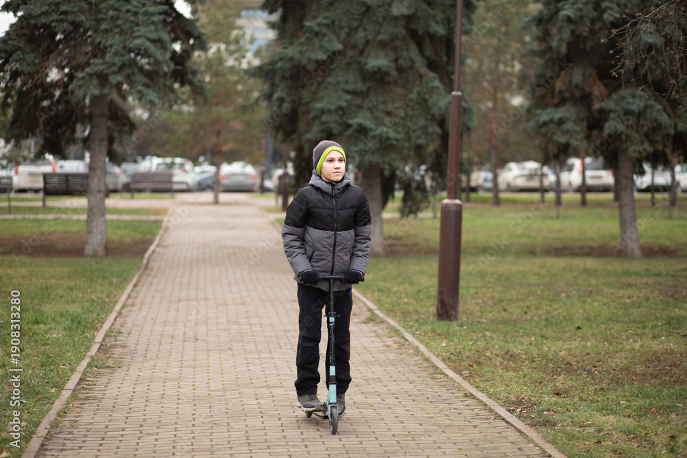 Fototapeta premium A child in a gray hooded jacket and black pants rides a blue kick scooter on a paved park path surrounded by trees and green grass.