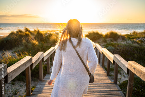 Beautiful middle-aged woman tourist walking down stairs overlooking sandy beach on sunset in spring. Back view
