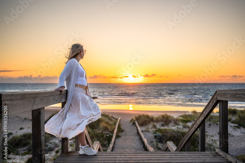 Algarve coast in Portugal. Beautiful mature woman tourist standing on wooden stairs overlooking sandy beach on sunset in spring. Side view	