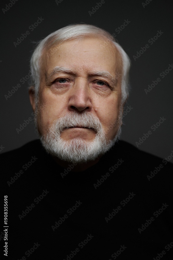 Fototapeta premium Older man with white beard looks directly at the camera in a dark setting