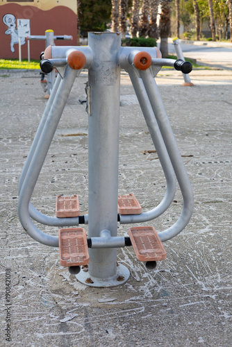 Vertical image of an outdoor fitness machine with pedals and handles installed in a public workout area. The equipment is designed for full-body exercise combining leg and upper body training.