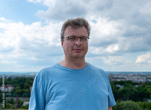 Close up portrait of a middle aged man wearing glasses and a blue t shirt on krakus mound overlooking krakow. Soft natural light, overcast cumulus sky, relaxed expression, urban skyline blur, and trav