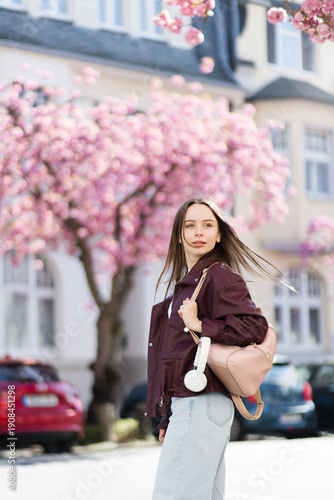 Young woman walking in spring city by cherry blossoms. Girl with backpack enjoying blooming trees, pink flowers. Urban lifestyle, cozy town. Casual street style.