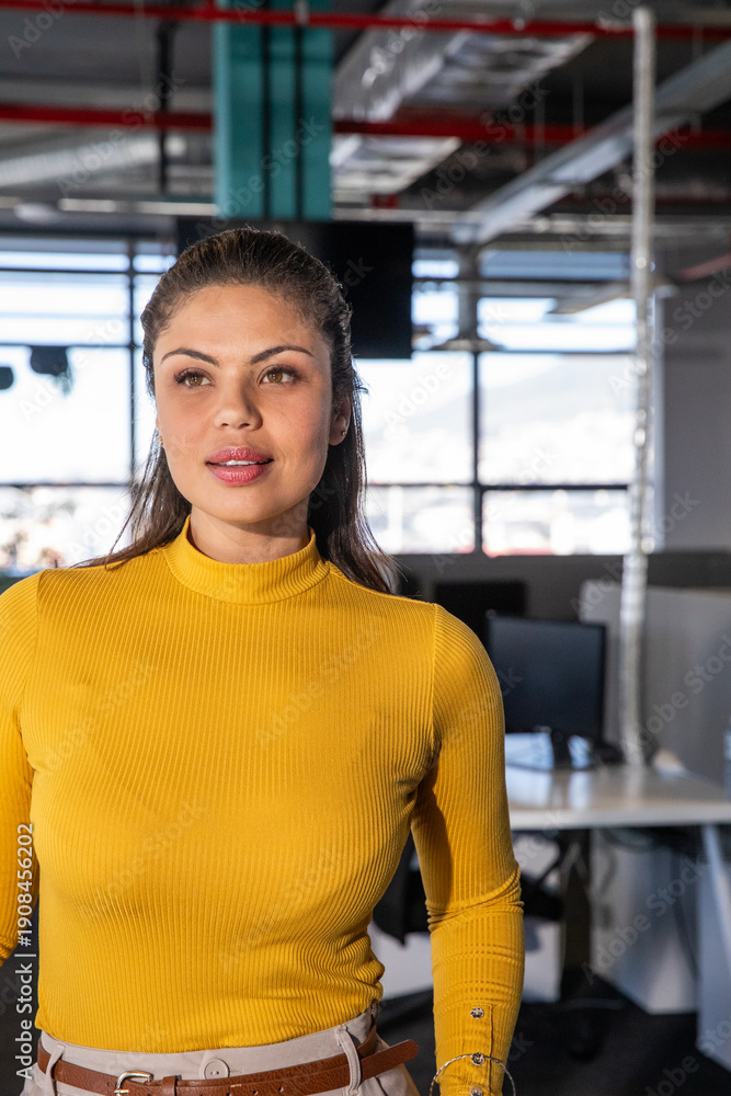 Fototapeta premium Woman standing, glancing right of camera in office with windows and monitors, wearing mustard top
