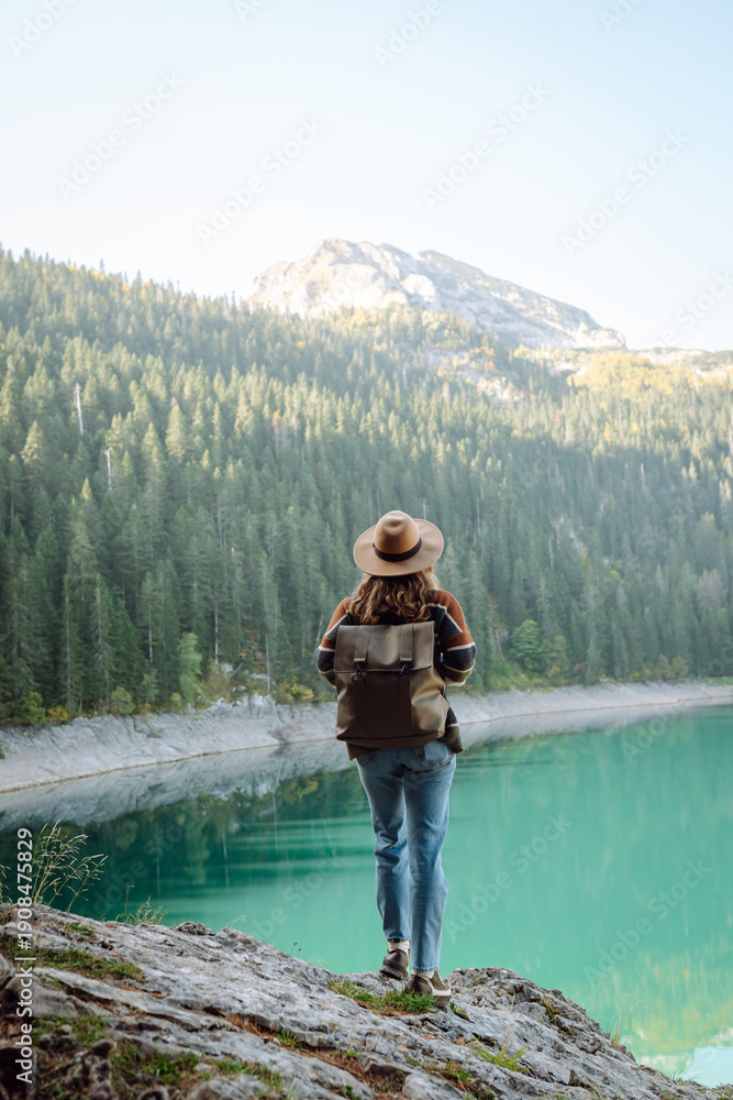 Naklejka premium A female traveler, wearing a hat and carrying a backpack, enjoys nature while sitting on a rock by a lake. A beautiful woman admires the mountain scenery at sunset. Concepts of adventure and freedom.
