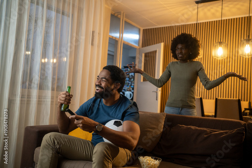 Unhappy wife watching husband ignore her while he drinks beer and watches TV