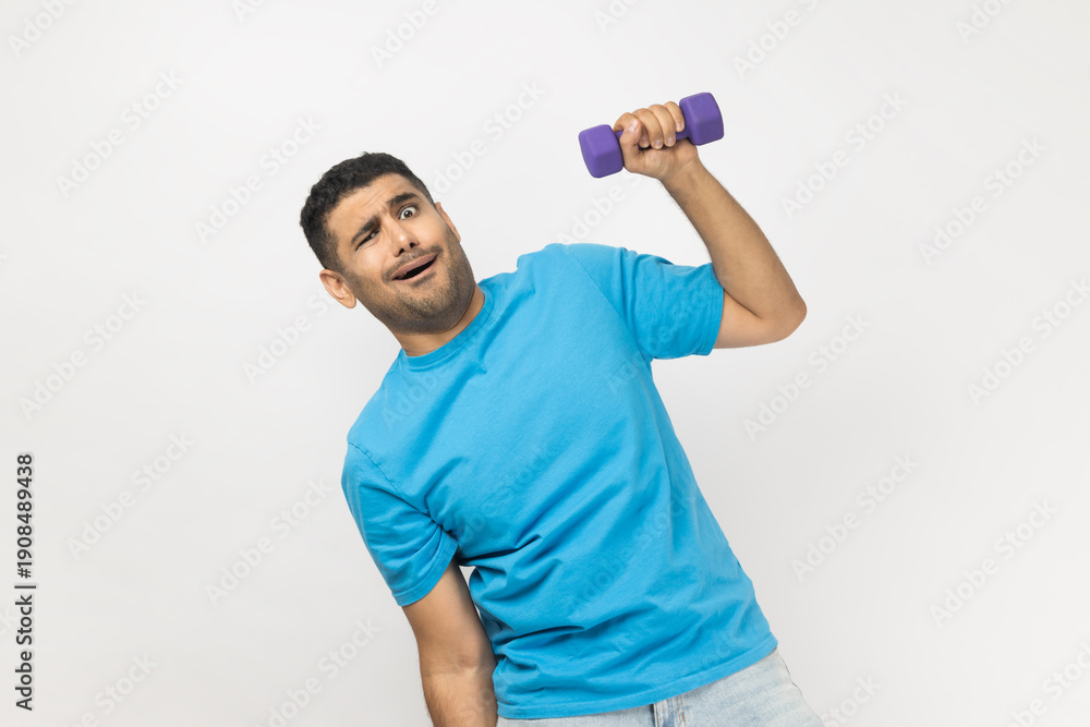 © khosrork - Portrait of funny playful unshaven man wearing blue T- shirt standing raised arm with dumbbell, pretended he raising heavy barbel. Indoor studio shot isolated on gray background.