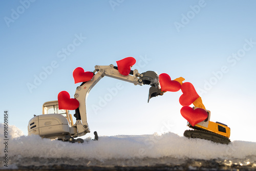 Two toy excavators reach out to each other in the snow with hearts on their arrows against a sky background. Concept: Love, construction business, romance. Business card for Valentine's Day