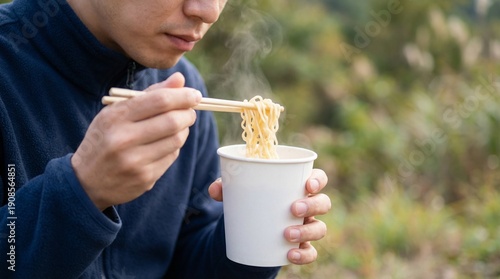 Person eating instant noodles from paper cup, wooden chopsticks lifting steaming noodles outdoors