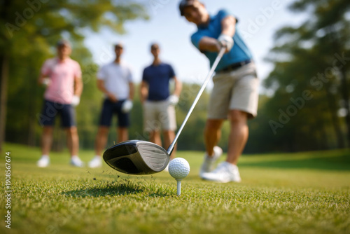 Golf player teeing off with a driver, hitting a golf ball on a tee with grass flying, surrounded by three blurred players in the background during an outdoor game on a golf course