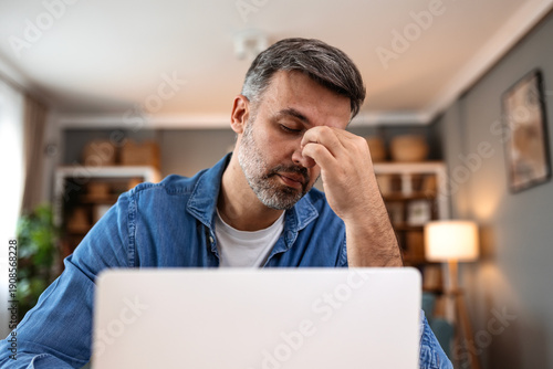 Tired man suffering from headache and eye strain while working on laptop. Healthcare and medicine concept. Stressed male professional holding bridge of nose with closed eyes at home office.