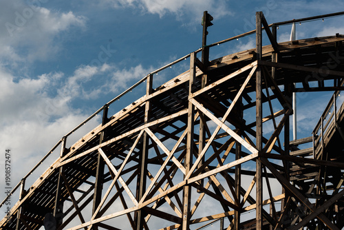 Close-up view of a towering wooden roller coaster structure with its intricate beams and tracks against a partly cloudy blue sky. Small lights line the railing