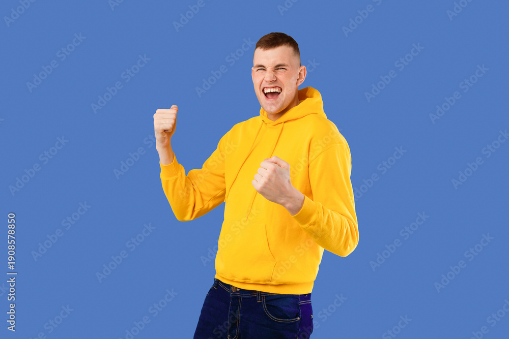 Naklejka premium Overjoyed caucasian man shouting and shaking fists, celebrating big success over blue studio background, free space. Celebration of victory, joy emotion concept