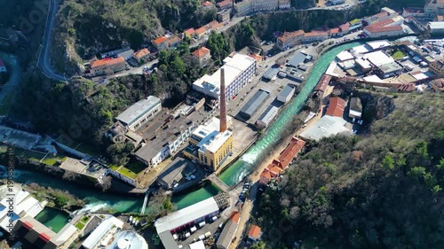 Aerial view of the Rijeka River winding through a valley, flanked by historic industrial buildings and residential areas under bright midday sun.