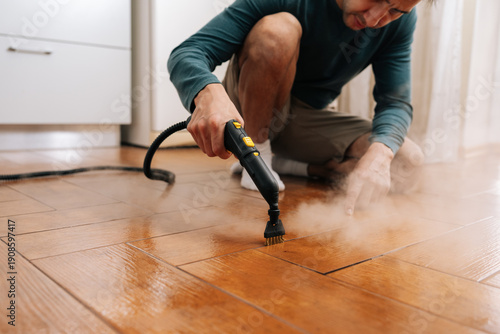 Fotografia Focused man steaming and scrubbing herringbone wood-look tile floor with brush attachment, removing grime and sanitizing grout for a fresh, spotless, healthy home environment