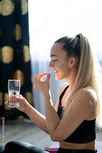 Fit woman taking vitamin pill with glass of water after workout at home, healthy lifestyle and wellness concept, morning routine in natural light.

