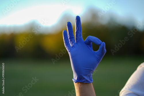Blue medical glove hand showing OK sign outdoors, healthcare approval gesture