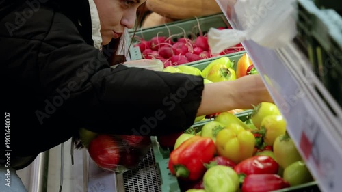 Young woman selecting vibrant bell peppers in a supermarket's vegetable aisle. She fills a plastic bag with her carefully chosen produce
