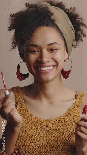 Young stylish african american lady demonstrating red lipgloss, choosing best makeup cosmetics, beige studio background