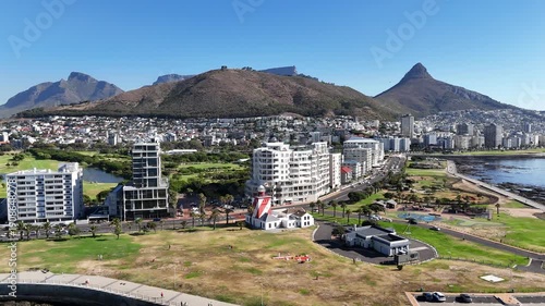 Drone orbits left in ultra wide shot of Green Point with Signal Hill, Lion's Head, and Table Mountain in the distance on a sunny day in Cape Town, South Africa
