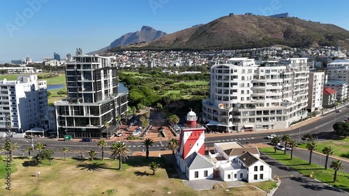 Drone flies backwards over red and white lighthouse in Green Point with view of Signal Hill on a sunny day in Cape Town, South Africa